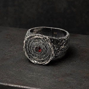 Puede incluir: Un anillo de plata con un acabado oxidado oscuro y muy texturizado, que presenta una cara circular con patrones concéntricos similares a los anillos de un árbol. Una pequeña piedra preciosa roja oscura está engastada en el centro. Marcado 925.