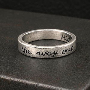 May include: A silver-toned metal ring with a distressed, weathered finish. The exterior band features the engraved black text "the way out" in a cursive script, while a partial word "Hid" is visible on the inner surface of the band.
