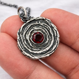 Op de afbeelding: Een zilveren hangerketting met een cirkelvormig ontwerp. De hanger heeft een getextureerd, gelaagd patroon rondom een ​​ronde, dieprode edelsteen. De ketting is ook zilverkleurig. De hanger heeft een diameter van ongeveer 2,5 cm.