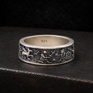May include: A silver ring with a dark, oxidized finish featuring embossed figures of a knight, a snail, and a deer. The interior of the band is clearly stamped with the number 925, indicating the sterling silver composition of the jewelry piece.