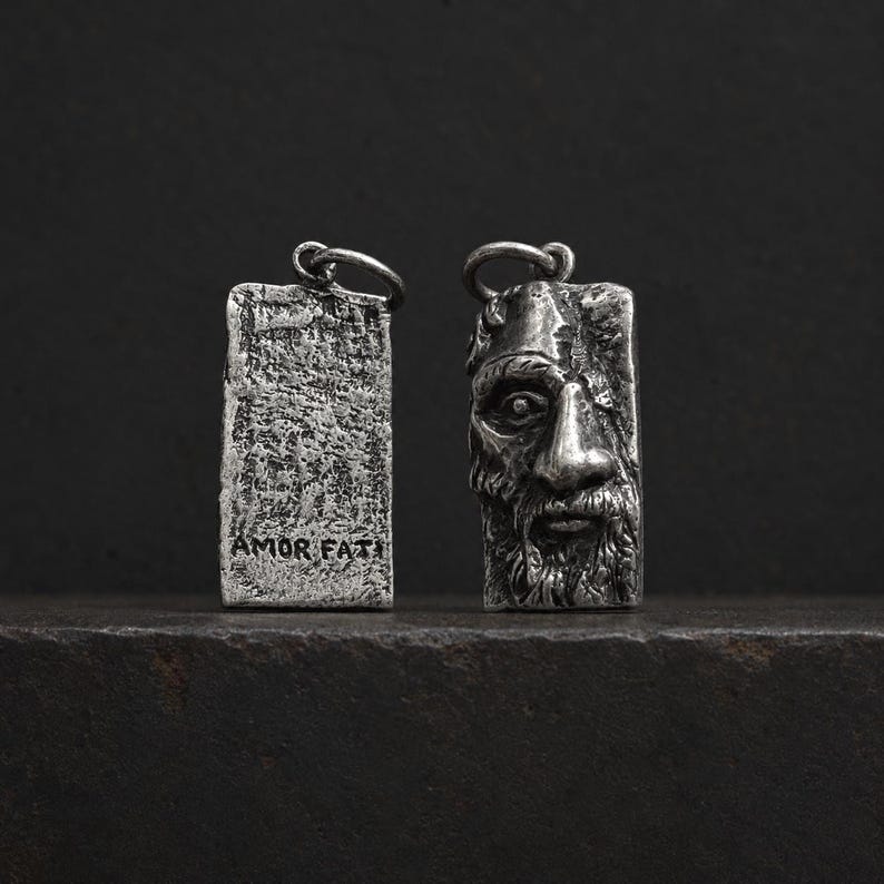 May include: Two rectangular silver-toned metal pendants with a weathered finish. One pendant features a sculpted face with a beard and mustache, while the other displays a textured surface with the engraved text AMOR FATI at the bottom.