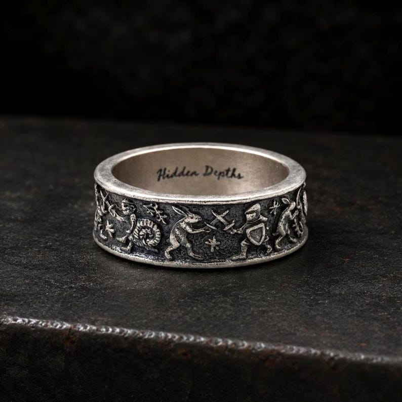 May include: A silver-toned metal ring featuring an embossed relief of rabbits and a knight engaged in a sword fight. The interior of the band is engraved with the text Hidden Depths in a cursive script, set against a dark, textured background.