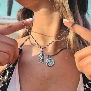 May include: A silver-toned necklace featuring two distinct pendants hanging from a dark metal chain. One pendant is a rectangular charm with a detailed relief of a bearded face, while the other is a circular yin-yang symbol with a hammered texture.