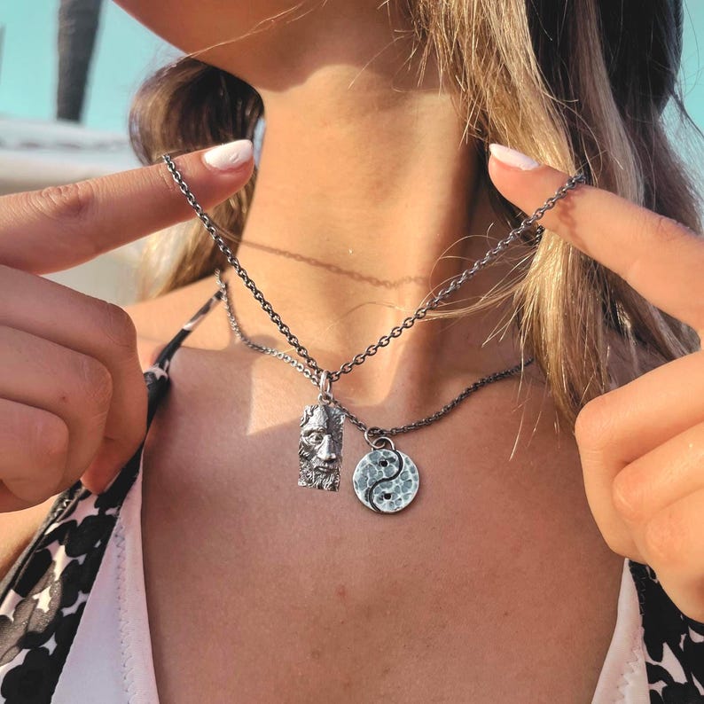 May include: A silver-toned necklace featuring two distinct pendants hanging from a dark metal chain. One pendant is a rectangular charm with a detailed relief of a bearded face, while the other is a circular yin-yang symbol with a hammered texture.