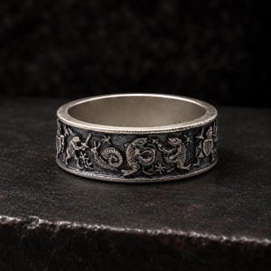 May include: A silver-toned metal ring featuring an embossed, detailed relief pattern around the exterior band. The design depicts a series of small, medieval-style figures, including a snail, various creatures, and armored knights holding shields.