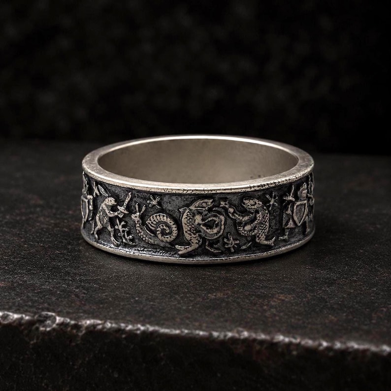 May include: A silver-toned metal ring featuring an embossed, detailed relief pattern around the exterior band. The design depicts a series of small, medieval-style figures, including a snail, various creatures, and armored knights holding shields.