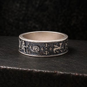 May include: A silver band ring featuring a dark, oxidized finish with intricate relief carvings. The design displays a series of figures including a knight with a sword, a snail, a cross-like emblem, and a winged creature in a repeating pattern.