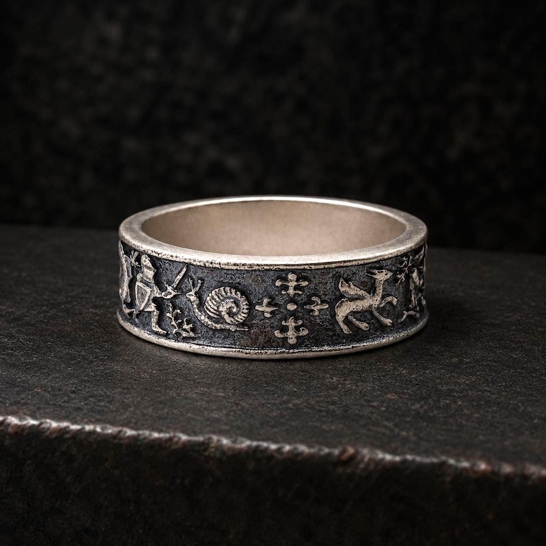 May include: A silver band ring featuring a dark, oxidized finish with intricate relief carvings. The design displays a series of figures including a knight with a sword, a snail, a cross-like emblem, and a winged creature in a repeating pattern.