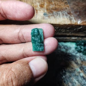 May include: A small, rectangular, translucent green gemstone carving featuring a detailed relief of a human face with a beard and mustache. The carving is held between fingers, showcasing the intricate facial features and the deep emerald hue.