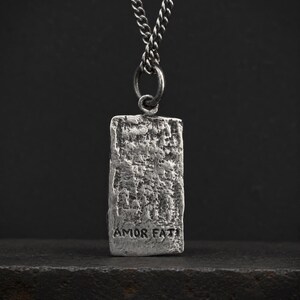 May include: A rectangular silver-toned pendant with a heavily textured, rugged surface suspended from a matching metal chain. The lower portion of the pendant features the stamped text AMOR FATI in a simple, dark, sans-serif capital letter font.