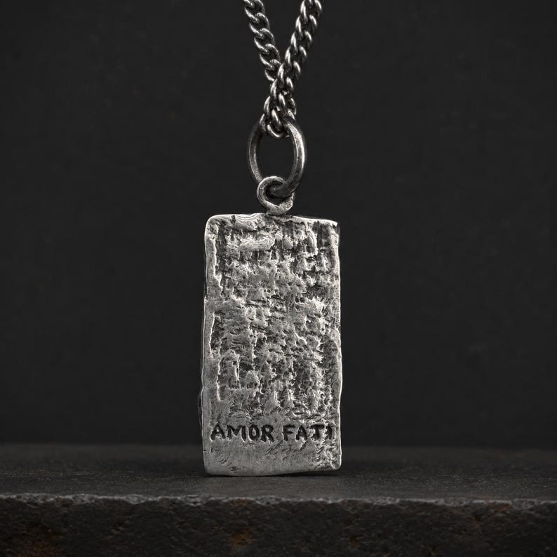 May include: A rectangular silver-toned pendant with a heavily textured, rugged surface suspended from a matching metal chain. The lower portion of the pendant features the stamped text AMOR FATI in a simple, dark, sans-serif capital letter font.