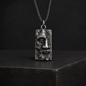 May include: A silver-toned rectangular pendant necklace featuring a detailed, high-relief sculptural face of a bearded individual. The pendant hangs from a matching silver-toned metal chain, showcasing an oxidized finish that highlights the facial features.