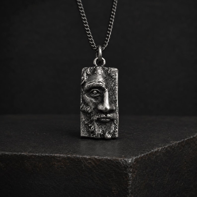 May include: A silver-toned rectangular pendant necklace featuring a detailed, high-relief sculptural face of a bearded individual. The pendant hangs from a matching silver-toned metal chain, showcasing an oxidized finish that highlights the facial features.