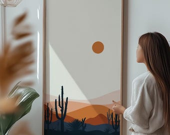 Può includere: Una stampa incorniciata di un paesaggio desertico con un grande sole arancione, cactus e montagne in tonalità di arancione, marrone e blu. La stampa è appesa a una parete bianca in un soggiorno moderno.