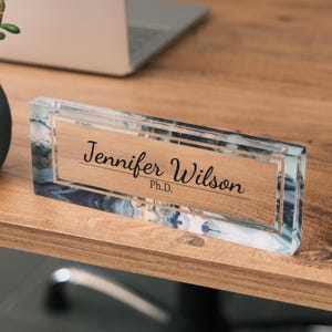 May include: A clear acrylic desk nameplate with a blue and white marbled design. The nameplate has a double border and the text "Jennifer Wilson Ph.D." is printed in black.