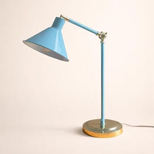 May include: A light blue desk lamp with a conical shade and adjustable arm. The lamp has a brass-coloured base and accents. The lamp is designed for desk use and has a power cord.