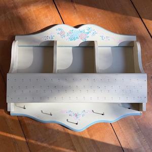 Peut inclure: Un organiseur mural en bois crème avec trois compartiments et une étagère inférieure avec des crochets. L'organisateur est décoré de motifs floraux roses et bleus et de rubans. Dimensions : environ 46 cm de large et 30 cm de haut.