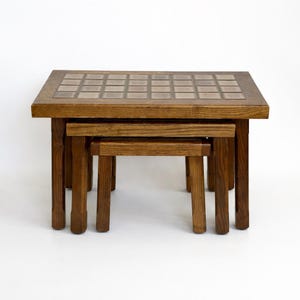 May include: A set of three wooden nesting tables. The largest table has a square top with a tiled surface. The tables are made of dark wood and have a simple, sturdy design. The smallest table fits neatly under the middle table.