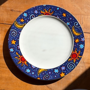 May include: A round ceramic dinner plate with a plain white center and a wide dark blue border. The border features a repeating pattern of yellow crescent moons, orange suns with rays, and various light blue and yellow stars and swirls.