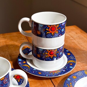 Op de afbeelding: Twee witte keramische mokken gestapeld op een bijpassende schotel, voorzien van een donkerblauwe band versierd met oranje en rode zonnen, gele sterren en blauwe krullen. Het ontwerp bevat kleine lichtblauwe bloemen en halve manen op de schotel.