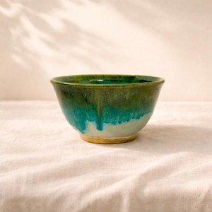 May include: A handmade ceramic bowl with a vibrant colour gradient. The bowl transitions from a deep green at the rim to a turquoise and white at the base. The bowl has a diameter of approximately 15 cm.