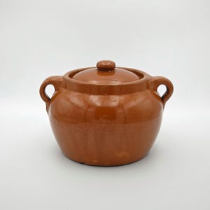 Könnte beinhalten: Ein brauner Keramiktopf mit Deckel und zwei Griffen. Der Topf hat eine abgerundete Form und eine glänzende Oberfläche. Der Deckel hat einen kleinen Knopf. Der Topf wird wahrscheinlich zum Kochen oder Servieren von Speisen verwendet.