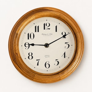 May include: A round wall clock with a light brown wooden frame and an off-white face. The clock features black serif numerals from 1 to 12, black hands, and the text Sterling & Noble Clock Company, registration, and Mfg. No. 9 printed on the face.