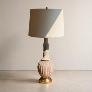 May include: A table lamp featuring a cylindrical off-white fabric shade, a spiraling textured stone-like stem, and a rounded, vertically ribbed base with a speckled finish. The lamp sits on a circular metallic gold-toned base at the bottom.