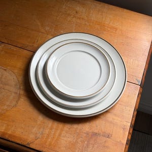 May include: Three white ceramic plates of varying sizes stacked on top of each other. Each plate has a thin, gold-colored rim. The plates are arranged on a wooden surface.