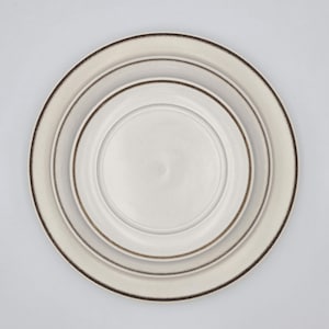 Può includere: Tre piatti in ceramica bianco sporco di varie dimensioni sono nidificati insieme. Il piatto più grande ha un bordo marrone scuro, il piatto centrale ha un bordo marrone e il piatto più piccolo è semplice. I piatti sono disposti a cerchio.