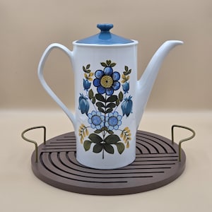 Peut inclure: Cafetière en céramique blanche avec couvercle et anse bleus, décorée d'un motif floral bleu, jaune et vert. La cafetière est posée sur un plateau rond marron avec des poignées en laiton. Article de cuisine de style vintage.