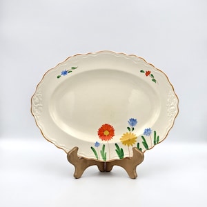 May include: Cream-coloured oval serving platter with a scalloped edge and a brown rim. The platter is decorated with colourful hand-painted flowers in red, yellow, and blue. The platter is displayed on a wooden stand.