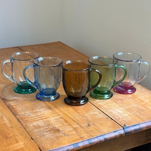 May include: Five clear glass mugs with colored bases and handles are arranged on a wooden table. The bases are green, blue, brown, green, and red. The mugs are ideal for serving hot beverages.