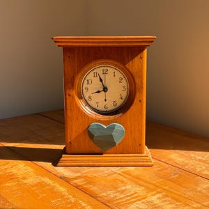 May include: A rectangular wooden mantel clock with a light brown finish. The clock face is circular with black numerals and hands. Below the clock face, a faceted, dark teal heart-shaped ornament is attached to the front of the wooden frame.