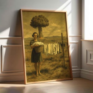Könnte beinhalten: Ein gerahmter Vintage-Druck zeigt eine lächelnde Frau, die einen Wäschekorb hält. Kleidung hängt auf einer Wäscheleine in einem Feld, mit einer Atompilzwolke im Hintergrund. Das Kunstwerk hat einen Sepia-Ton und ist in hellem Holz gerahmt.