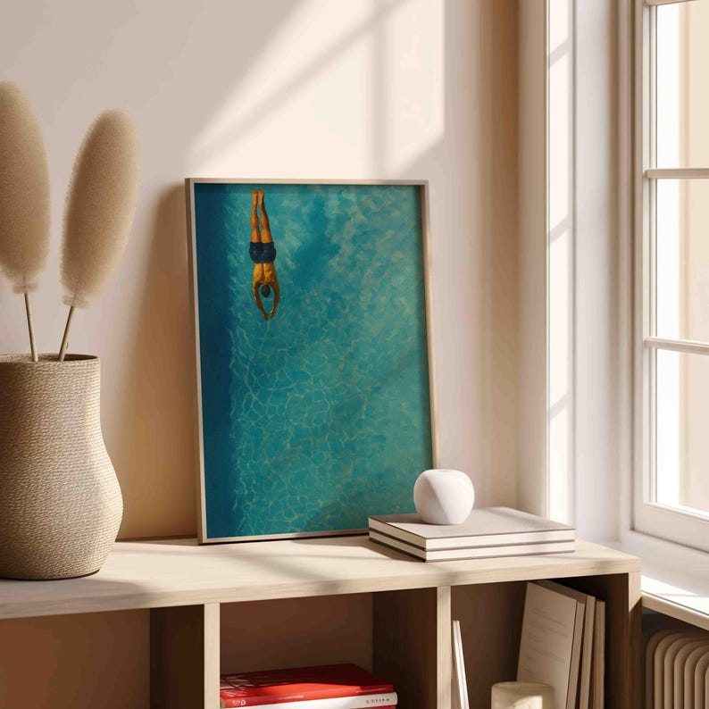 May include: Framed art print featuring a person diving into a turquoise pool. The artwork has a gold frame and is displayed on a light-colored wooden shelf. A decorative vase with dried plants and a stack of books are also on the shelf.