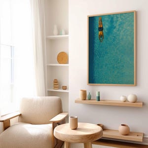 May include: A framed art print depicts a person diving into a turquoise pool. The artwork is in a light wood frame and hangs on a white wall. The room features a neutral color palette with wooden furniture and decorative vases.