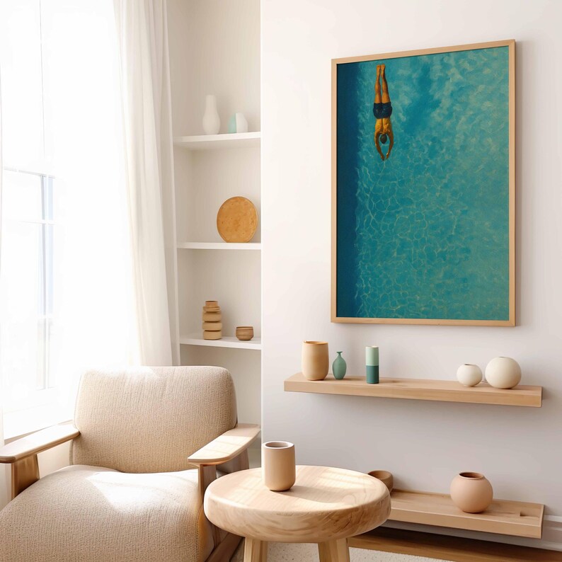 May include: A framed art print depicts a person diving into a turquoise pool. The artwork is in a light wood frame and hangs on a white wall. The room features a neutral color palette with wooden furniture and decorative vases.