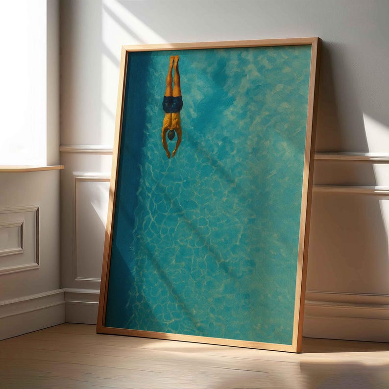 May include: A framed art print depicts a person diving headfirst into a turquoise pool. The person is wearing dark blue swim trunks. The artwork is framed in a light brown wooden frame and leans against a white wall.
