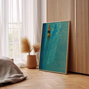 May include: A framed art print featuring a person diving into a turquoise pool. The artwork is displayed on a wooden floor, leaning against a wooden slatted wall. A woven basket with dried plants sits near a window with white curtains.