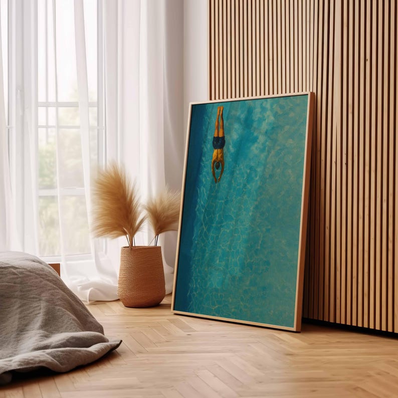 May include: A framed art print featuring a person diving into a turquoise pool. The artwork is displayed on a wooden floor, leaning against a wooden slatted wall. A woven basket with dried plants sits near a window with white curtains.