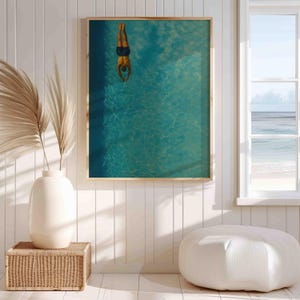 May include: A framed art print depicts a person diving headfirst into a turquoise pool. The artwork is set in a bright room with a window overlooking the ocean. A white vase with dried plants and a woven basket are in the foreground.