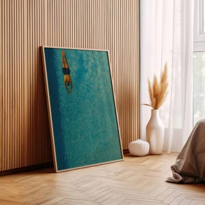 May include: A framed art print depicts a person diving into a turquoise pool. The artwork is displayed against a wooden wall and floor, with a white vase and decorative plant in the background. The print has a light wood frame.
