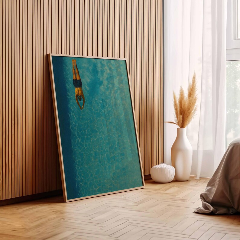 May include: A framed art print depicts a person diving into a turquoise pool. The artwork is displayed against a wooden wall and floor, with a white vase and decorative plant in the background. The print has a light wood frame.
