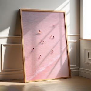 May include: A framed art print depicts a serene beach scene. The artwork showcases a pink ocean with several figures swimming and floating in the water. A person is in a pink inflatable ring. The frame is a light brown color.