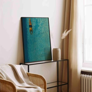 May include: A framed art print depicts a person diving into turquoise water. The artwork is displayed on a black metal and glass console table, with a woven chair and beige throw blanket in the foreground. A beige vase with dried plants sits on the table.