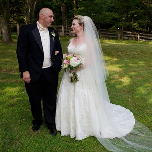 May include: A bride and groom stand in a grassy field, the bride wearing a white wedding dress with a long veil and a bouquet of pink and white flowers. The groom is wearing a black tuxedo.