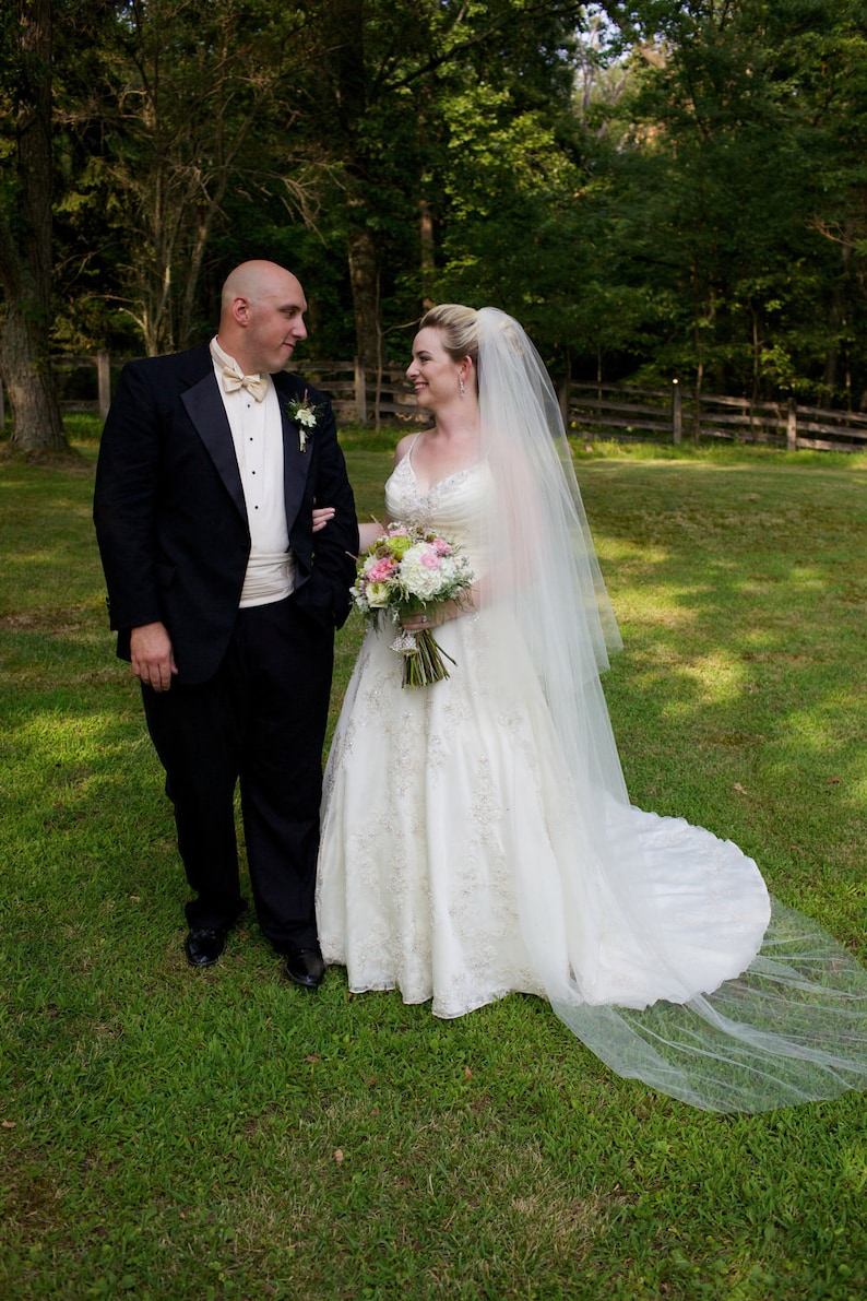 May include: A bride and groom stand in a grassy field, the bride wearing a white wedding dress with a long veil and a bouquet of pink and white flowers. The groom is wearing a black tuxedo.