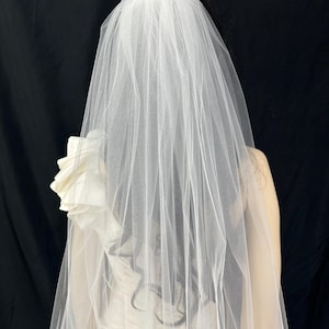 K&ouml;nnte beinhalten: Ein wei&szlig;er Brautschleier aus transparentem Stoff f&auml;llt &uuml;ber den R&uuml;cken. Der Schleier ist an einer Frisur mit dunklem Haar befestigt. Ein wei&szlig;es Kleid mit floralem Akzent ist sichtbar.