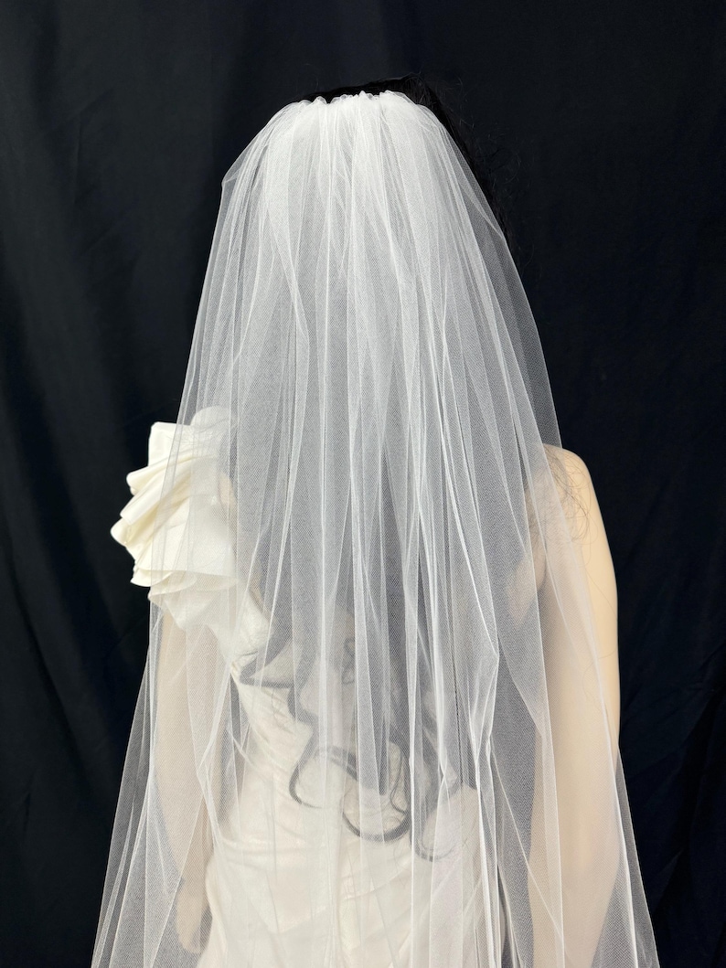 K&ouml;nnte beinhalten: Ein wei&szlig;er Brautschleier aus transparentem Stoff f&auml;llt &uuml;ber den R&uuml;cken. Der Schleier ist an einer Frisur mit dunklem Haar befestigt. Ein wei&szlig;es Kleid mit floralem Akzent ist sichtbar.